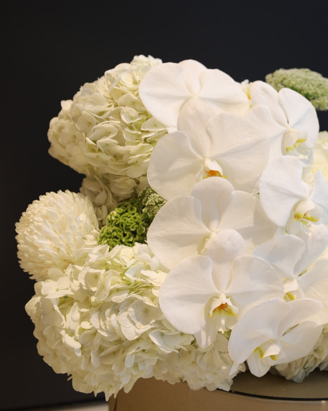 A close-up of a floral arrangement featuring white orchids, cream hydrangeas, and small green filler blooms against a dark background.