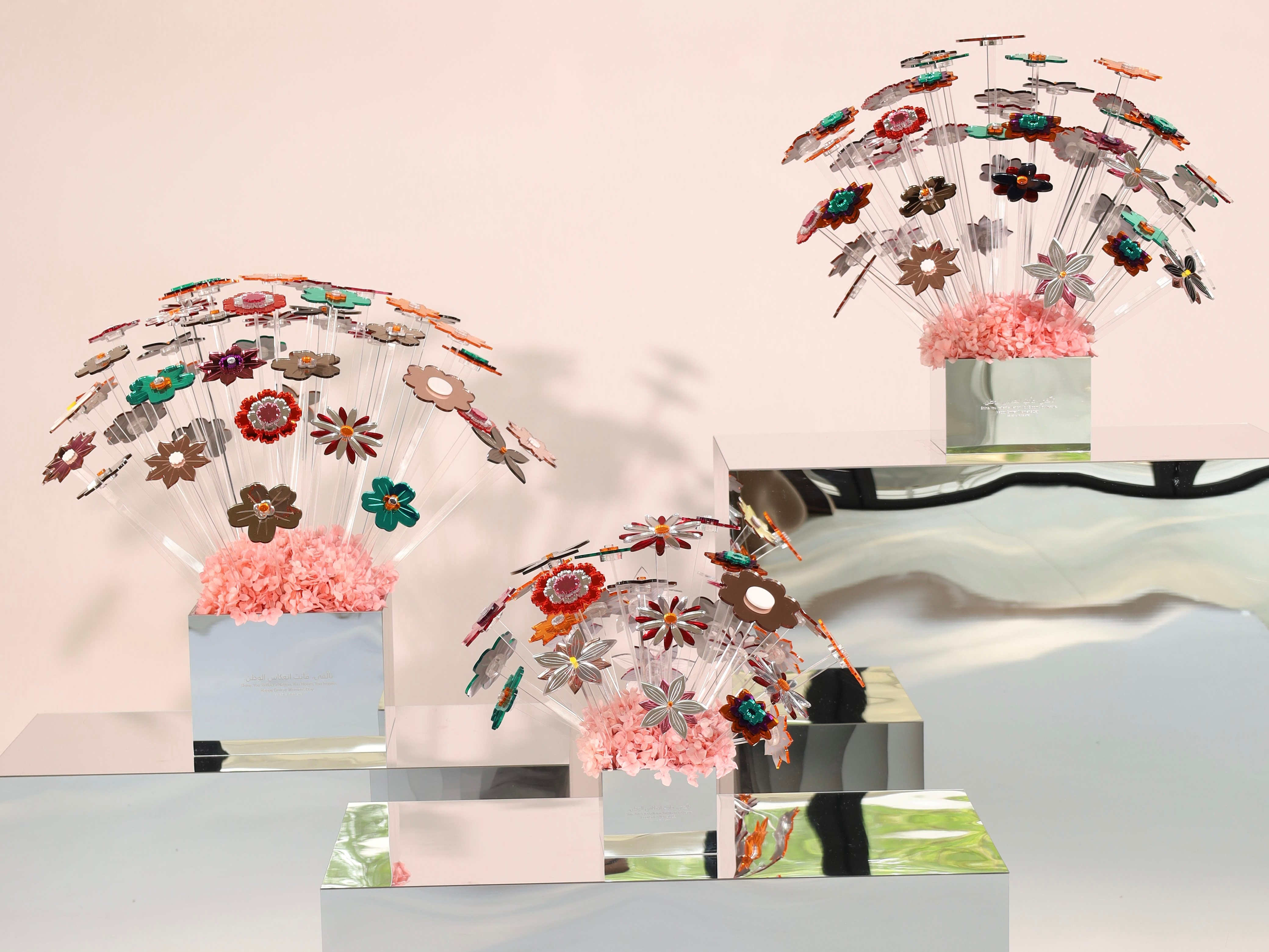 Three decorative floral arrangements in clear domed containers with pink paper filler, displayed on mirrored white pedestals against a pale pink background.